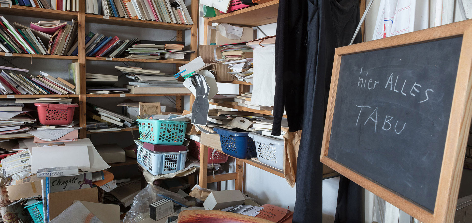 unordentliches Zimmer, voller Bücher, Zettel, Kisten; im Vordergrund Tafel mit der Aufschrift "alles tabu"
