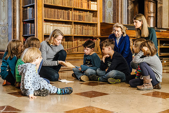 Eine Gruppe Kinder und drei Frauen hocken am Boden des Prunksaals und betrachten ein Buch