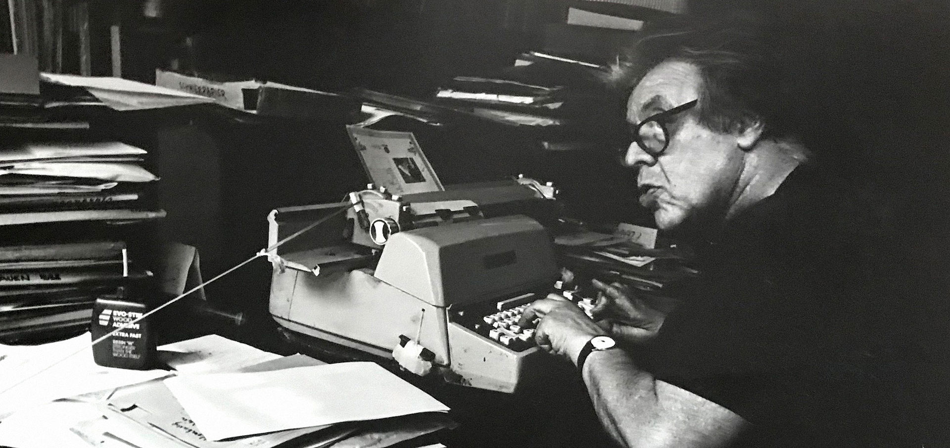 Schwarz-weiß Foto von Mann, der an überfülltem Schreibtisch and einer Schreibmaschine sitzt