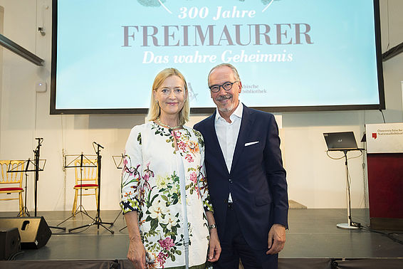 Ein Mann im Anzug und eine Frau stehen vor einer Bühne. Hinter ihnen sieht man eine Leinwand, auf die die Worte "Freimaurer - Das wahre Geheimnis" projiziert werden.