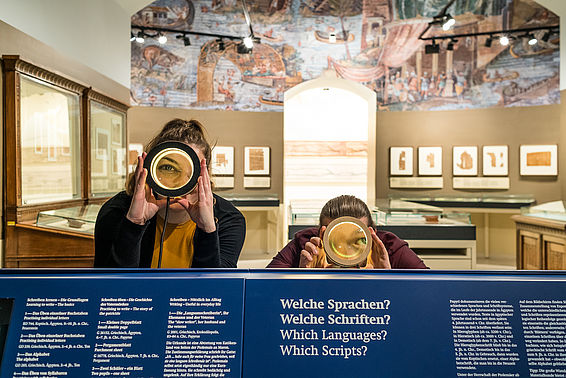 BesucherInnen schauen durch Vergrößerungsgläser im Papyrusmuseum, Raum mit Wandmosaik im Hintergrund