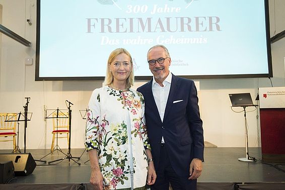 Blonde Frau im Blumenblazer und ein Mann im schwarzen Anzug vor einem Screen mit dem Design der Freimaurer Ausstellung