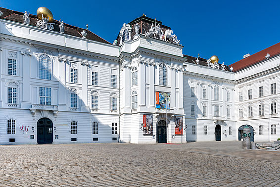 Foto eines dreistöckigen, weißen Gebäudes mit Statuen und goldenen Kugeln am Dach, davor eine leerer Platz