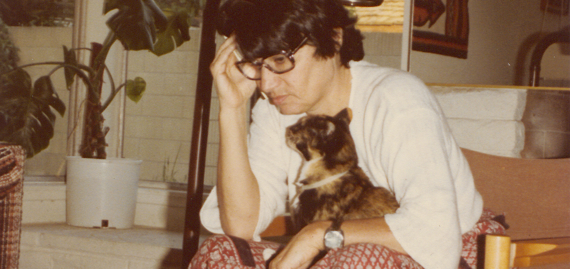 Älteres Foto von einer Frau mit Brille und schwarzen Haaren, die mit einer Katze auf dem Schoß an einem Couchtisch sitzt und nachdenkt.