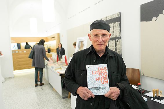 Schwarz gekleideter Mann mit schwarzer Kopfbedeckung (Bodo Hell) mit dem Museumskatalog des Literaturmuseums