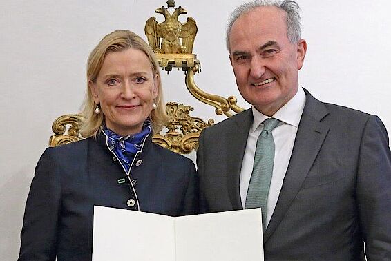 Eine blonde Frau und ein kahlköpfiger Mann im Anzug halten eine Urkundende