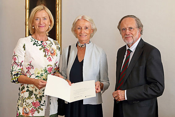 Eine Frau überreicht einer anderen Frau eine Urkunde. Daneben steht ein Mann im Anzug.