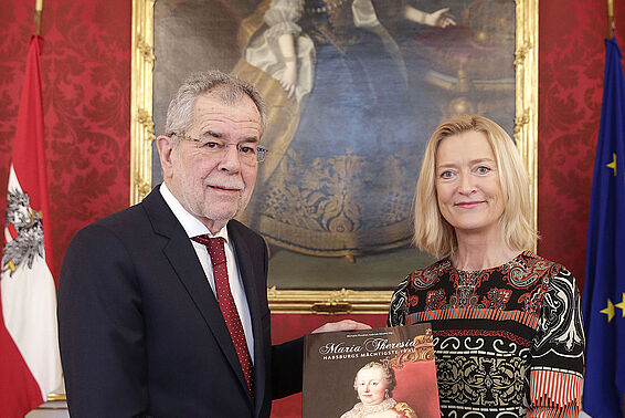 Ein Mann und eine Frau stehen vor einem Gemälde. Die Frau überreicht dem Mann einen Katalog.