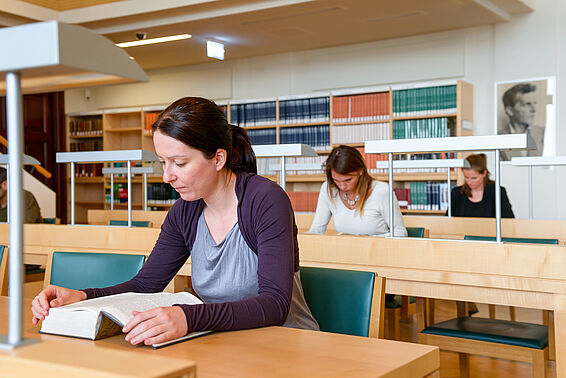 Drei Frauen sitzen an Schreibtischen in einem Saal mit Bücherregalen und lesen in Büchern