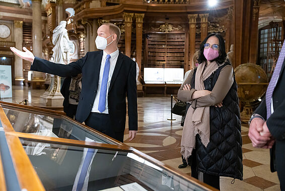 Einige Personen in eleganter Kleidung stehen in einem Marmorsaal vor Vitrinen