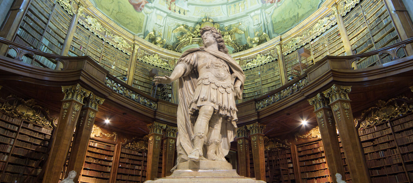 Prunksaal Foto aufschauend zu Deckenfresko und Statue, im Hintergrund Bücherregale