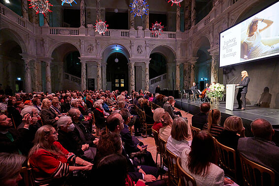 Volles Publikum vor Bühne in prunkvollem Saal.