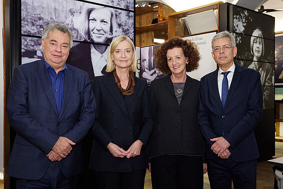 Zwei Frauen und Männer posieren vor Museumsregalen mit schwarz-weißen Fotos.