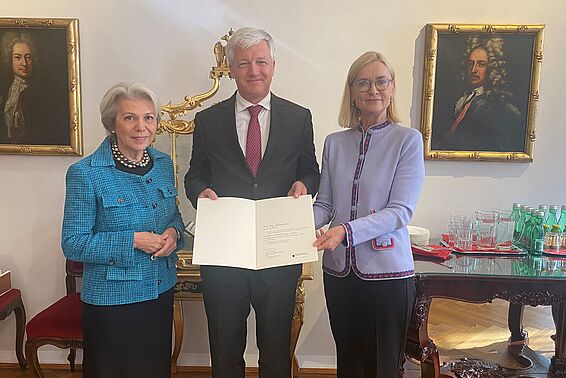 Ein weißhaariger Mann im schwarzen Anzug, links eine weißhaarige Frau im blauen Kostüm, rechts einer blonden Frau im fliederfarbenen Sacko