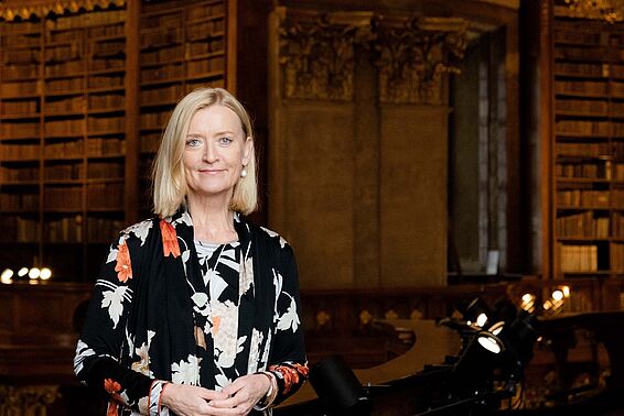 Porträtfoto von einer blonden Frau in dunkler, gemusterter Bluse vor Bücherregalen