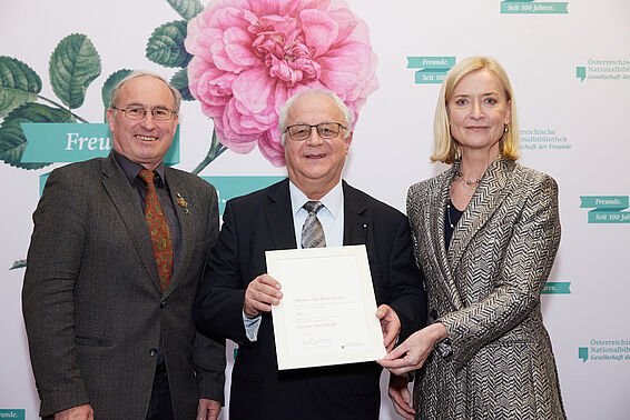 Zwei Männer und eine Frau halten eine Urkunde vor einer Logowand mit rosa Rose.