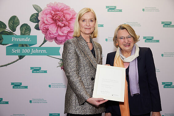 Zwei Frauen halten eine Urkunde vor einer Logowand mit rosa Rose.