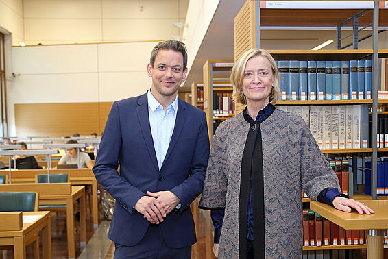 Ein Mann im Anzug und eine Frau stehen in einem Lesesaal voller Bücherregale und Schreibtischen.