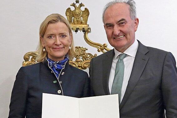Eine blonde Frau und ein kahlköpfiger Mann im Anzug halten eine Urkundende