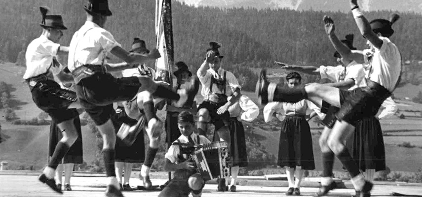 Tänzer und Tänzerinnen in Tracht tanzen im Kreis vor Bergkulisse, schwarz-weiß