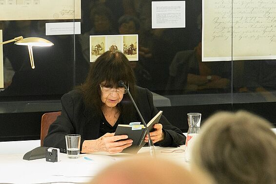 Schwarz gekleidete Dame mit schwarzen Haaren (Friederike Mayröcker) liest vor Publikum aus einem Buch