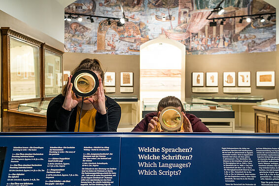 BesucherInnen schauen durch Vergrößerungsgläser im Papyrusmuseum, Raum mit Wandmosaik im Hintergrund