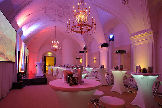 Pink beleuchteter Raum mit Stehtischen, Bühne und Kronleuchter