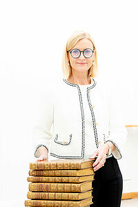 Foto einer Frau mit weißer Jacke, die hinter einem Stapel alter Bücher steht
