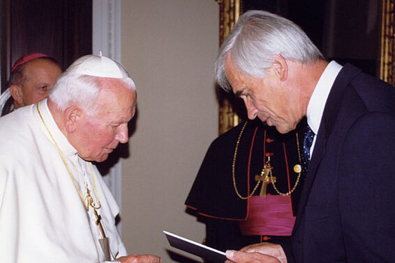 Ein weißhaariger Mann im schwarzen Anzug überreicht Papst Johannes Paul II eine Urkunde