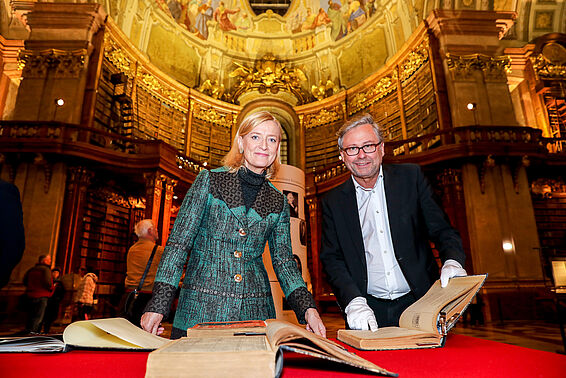 Frau und Mann posieren mit alten Buchen auf einem Tisch, im Hintergrund sieht man das Deckenfresko