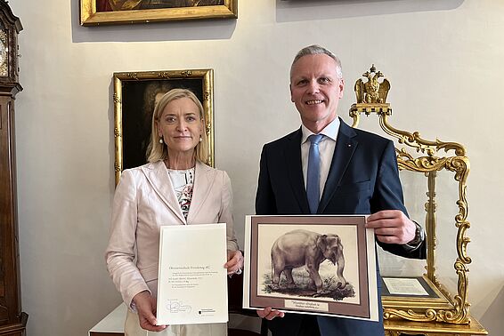 Blonde Frau mit Urkunde in der Hand und grauhaariger Mann mit Elefantenbild in der Hand