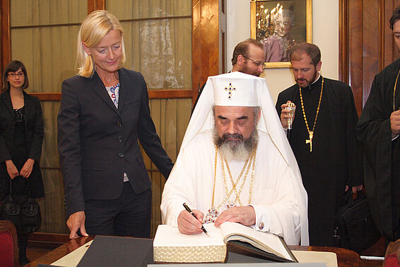 Frau steht neben Geistlichem in weißen Gewändern, der in einem Buch unterschreibt