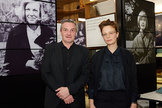 Eine Frau und ein Mann posieren vor Museumsregalen mit schwarz-weißen Fotos.