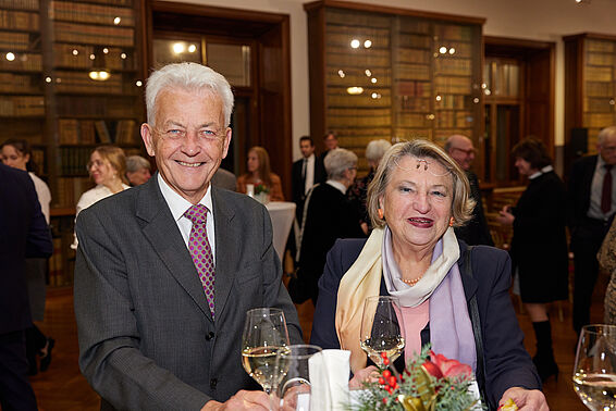 Ein Mann und eine Frau trinken Wein an einem Stehtisch, im Hintergrund sieht man eine Menge und Bücherregale an den Wänden.