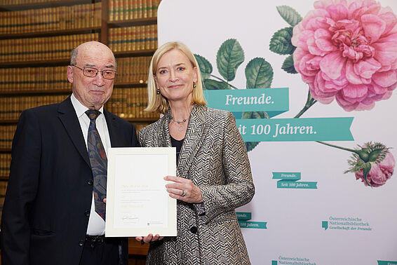 Ein Mann und eine Frau halten eine Urkunde vor einer Logowand mit rosa Rose.