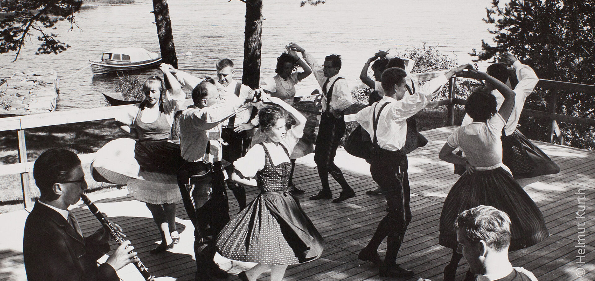 Schwarz-weißes Foto von einer Gruppe von Tanzpaaren in Tracht, dahinter ein See.