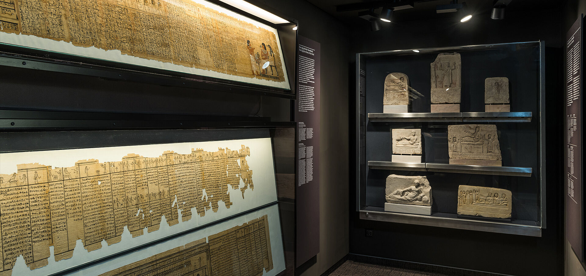 Foto von antiken Steintafeln sowie einem langen Stück Papyrus, beide hinter Glas