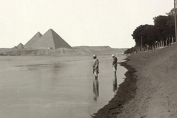 Schwarz-weißes Foto von zwei Personen die am Ufer im Wasser stehen, im Hintergrund Pyramiden