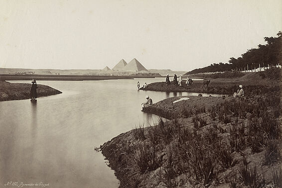 Menschen am Flussufer, im Hintergrund Pyramiden.