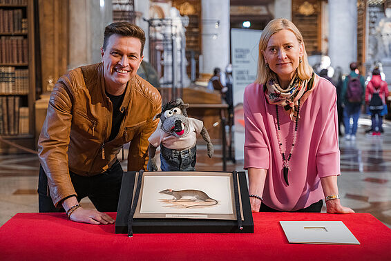 Mann, eine Ratten-Handpuppe und eine Frau posieren mit einer Ratten-Aquarellzeichnung