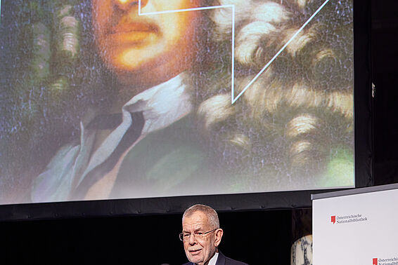 Grauhaariger Mann an Rednerpult vor Porträt von Mann mit gelockter Perücke