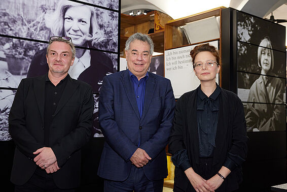 Eine Frau und zwei Männer posieren vor Museumsregalen mit schwarz-weißen Fotos.