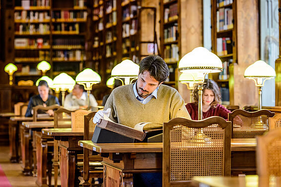 Ein Mann liest vorsichtig in einem alten Buch in einem Saal voller Schreibtische, Lampen und Bücherregalen