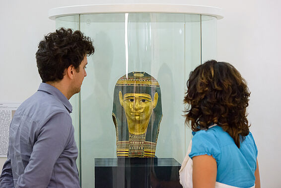 Zwei Personen sehen sich eine Mumienmaske in einer Glasvitrine an