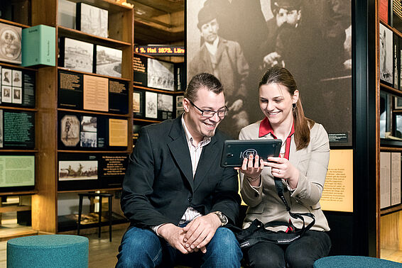 Ein Mann und eine Frau sehen sich in einem Museumsraum mit Regalen etwas auf einem Tablet an