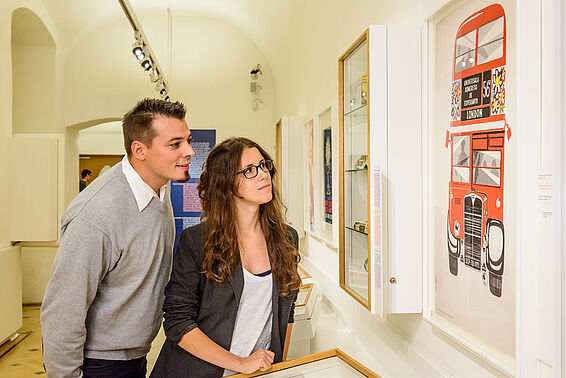Ein Mann und eine Frau sehen sich in einem Museumsraum ein Poster von einem roten Doppeldeckerbus an