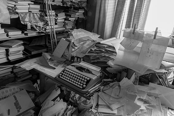 Schreibtisch und Schreibmaschine in einem Zimmer vollgestopft mit Büchern und Notizen, schwarz-weiß