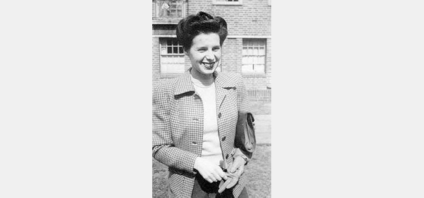 Frau mit karierter Jacke und Aktentasche unterm Arm, vor Backsteinhaus, schwarz-weiß