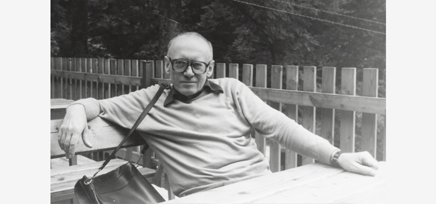 Schwarz-weißes Foto von einem Mann mit Brille und Pullover, der an einer Bank vor einem Zaun lehnt und in die Kamera blickt