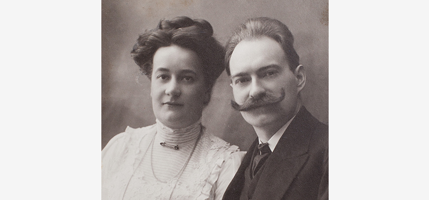 Schwarz-weißes Foto von einem Mann und einer Frau. Sie hat hochgesteckte, dunkel Haare und einen hellen Rollkragen. Er trägt einen gekräuselten Schnurrbart und einen dunklen Anzug.